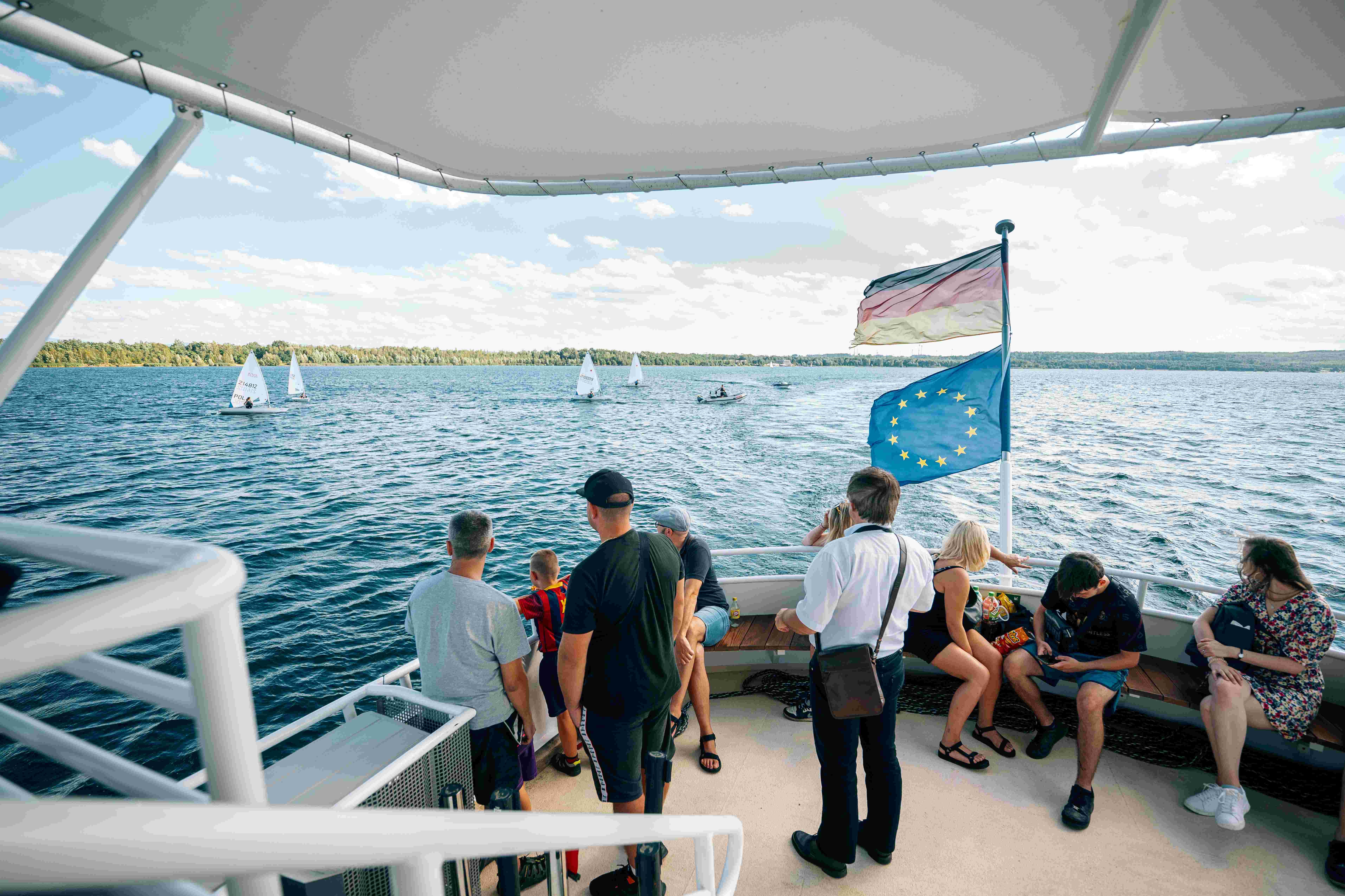 Passengers stand at the bow of a passenger ship and look out over a large lake.